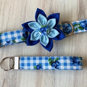 May include: A blue and white gingham dog collar with a blue flower charm and a matching lead. The collar and lead feature a blueberry print.