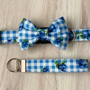 Op de afbeelding: Blauw-witte gingham hondenhalsband en strik met een blauwe bessenprint. De halsband heeft een zilveren gesp en de strik heeft een zwarte gesp.