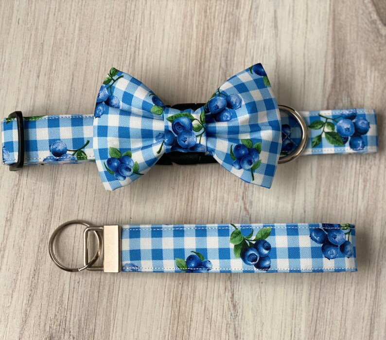 Op de afbeelding: Blauw-witte gingham hondenhalsband en strik met een blauwe bessenprint. De halsband heeft een zilveren gesp en de strik heeft een zwarte gesp.