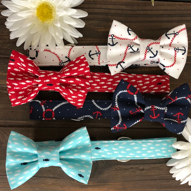 May include: A collection of dog collars with bow ties in various nautical patterns. Designs include anchors, fish, and rope motifs in red, white, and navy blue. The collars are displayed on a wooden surface with white flowers.