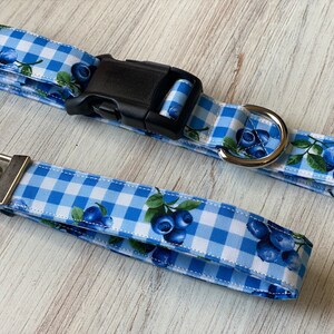 Op de afbeelding: Een blauwe en witte gingham hondenriem met een blauwe bessenprint en een bijpassende sleutelhanger. De riem heeft een zwarte plastic gesp en een zilveren metalen D-ring.