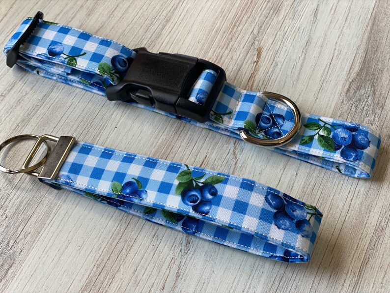Op de afbeelding: Een blauwe en witte gingham hondenriem met een blauwe bessenprint en een bijpassende sleutelhanger. De riem heeft een zwarte plastic gesp en een zilveren metalen D-ring.