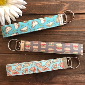 Op de afbeelding: Drie sleutelhangers met verschillende voedselontwerpen. De eerste sleutelhanger is blauw met een witte achtergrond en heeft taco's. De tweede sleutelhanger is grijs met een witte achtergrond en heeft hamburgers. De derde sleutelhanger is blauw met een witte achtergrond en heeft pizzapunten.