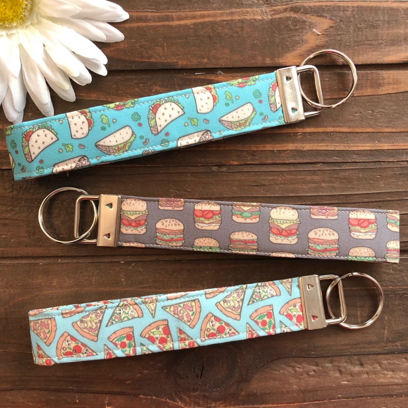 Op de afbeelding: Drie sleutelhangers met verschillende voedselontwerpen. De eerste sleutelhanger is blauw met een witte achtergrond en heeft taco's. De tweede sleutelhanger is grijs met een witte achtergrond en heeft hamburgers. De derde sleutelhanger is blauw met een witte achtergrond en heeft pizzapunten.