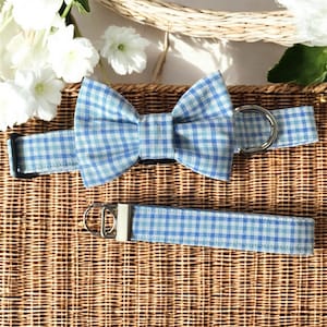 May include: A blue and white gingham dog collar and matching bow tie set. The collar has a black buckle and a silver metal ring. The bow tie is pre-tied. The set is displayed on a woven basket.