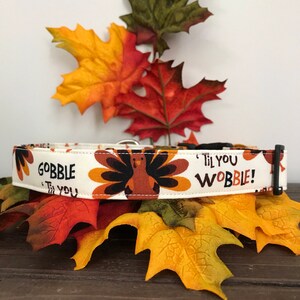 Może przedstawiać: Biały obroża dla psa z nadrukiem indyka i tekstem "Gobble 'Til You Wobble!" Obroża leży na tle jesiennych liści.