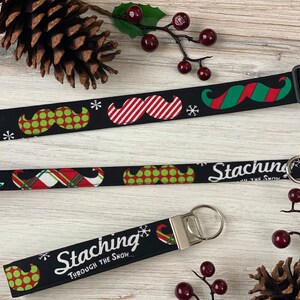 May include: Black and green Christmas-themed dog collar and leash with a repeating pattern of mustaches and snowflakes. The collar has a black buckle and the leash has a silver key ring. The text "Stacking Through The Snow..." is printed on the leash.