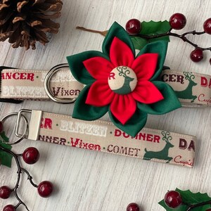 May include: A beige dog collar with a red and green poinsettia flower and a reindeer print. The collar has the words "Dancer, Blitzen, Vixen, Donner, Comet, Cupid" printed on it. The collar is paired with a matching leash.