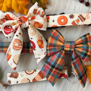 May include: Two dog collars with matching bow ties. One collar is white with a brown, orange, and white pattern of doughnuts, coffee cups, and cinnamon sticks. The other collar is a plaid pattern of orange, red, blue, and black. Both collars have matching bow ties.