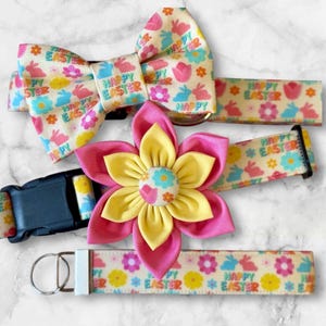 May include: A set of three dog accessories featuring a yellow and pink flower, a bow tie, and a collar. All three items are decorated with a white background and a repeating pattern of colorful flowers, bunnies, and the text "Happy Easter".