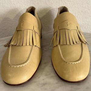 Puede incluir: Un par de mocasines de cuero amarillo claro con flecos. Los zapatos tienen una puntera redondeada, una costura decorativa y un peque&ntilde;o logotipo en la parte superior. Las suelas son de color marr&oacute;n oscuro.