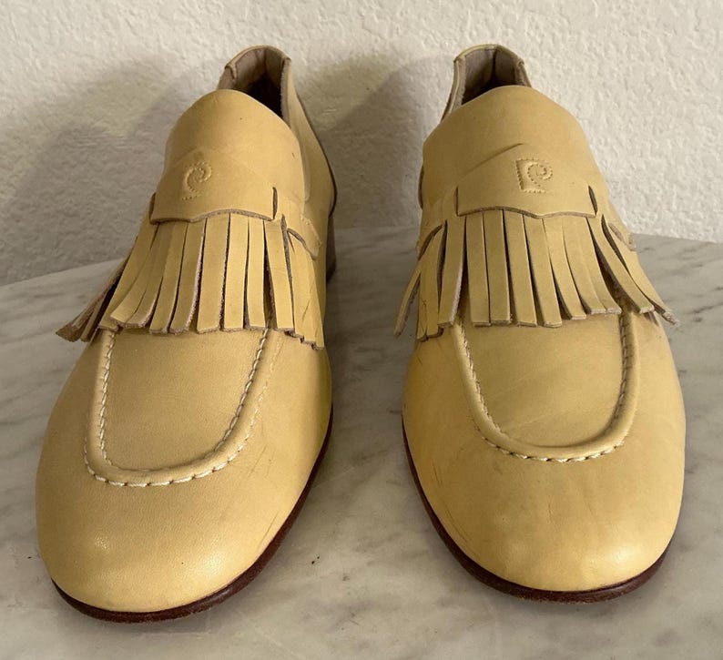 Puede incluir: Un par de mocasines de cuero amarillo claro con flecos. Los zapatos tienen una puntera redondeada, una costura decorativa y un peque&ntilde;o logotipo en la parte superior. Las suelas son de color marr&oacute;n oscuro.