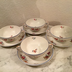 Może przedstawiać: Zestaw czterech białych porcelanowych misek do zupy z niebieską obwódką i kwiatowymi wzorami. Każda miska ma dwa uchwyty i jest dostarczana z dopasowanym spodkiem.