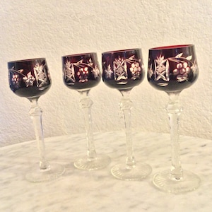 May include: Set of four clear glass stemmed wine glasses with a deep purple hue and intricate cut crystal designs. The glasses have a round bowl and a long, slender stem.
