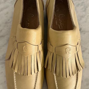Puede incluir: Un par de mocasines de cuero color beige claro con flecos. Los zapatos tienen una puntera redondeada y una correa decorativa con un logotipo. Los zapatos est&aacute;n marcados "Pierre Cardin" y "Made in Italy".