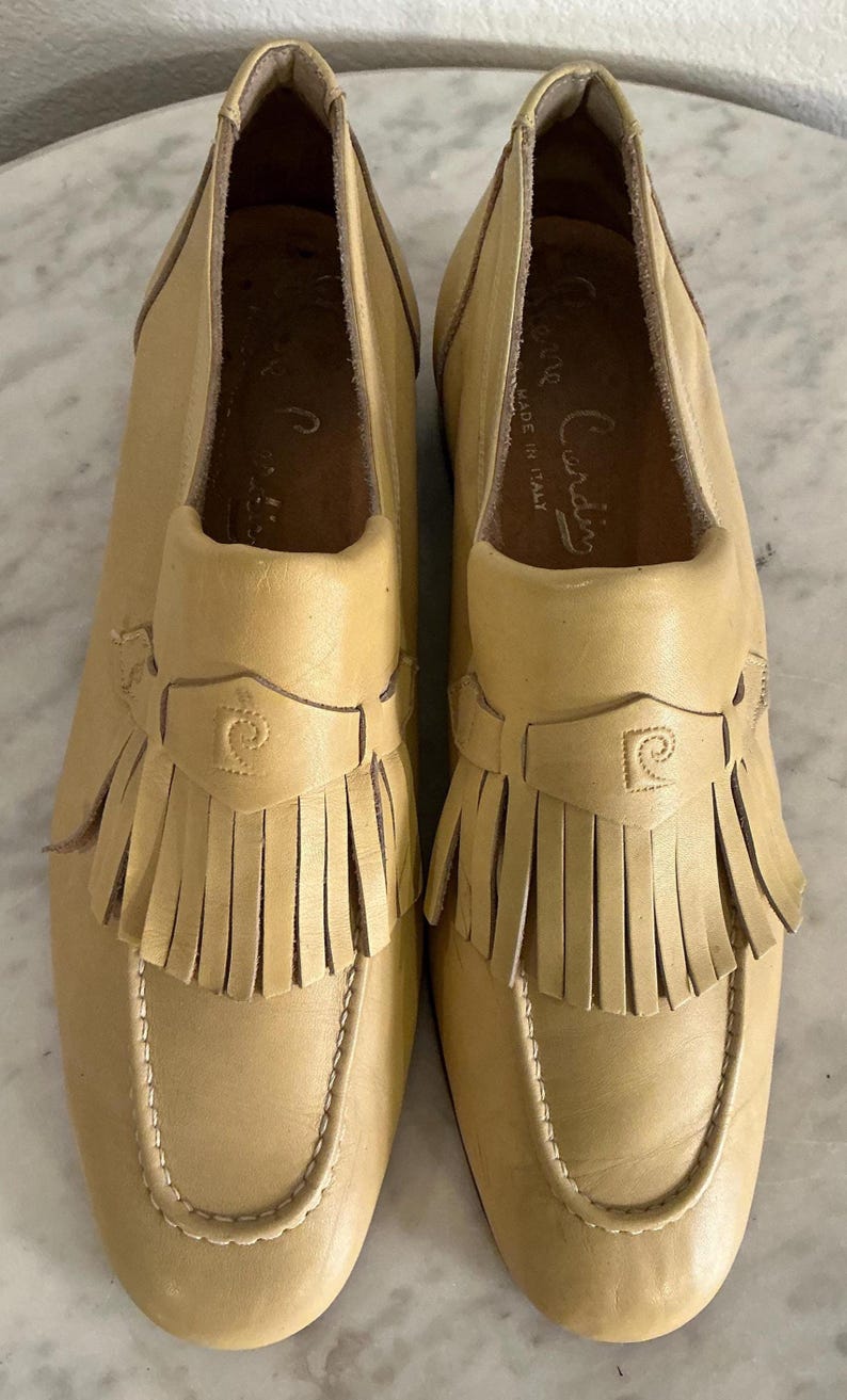 Puede incluir: Un par de mocasines de cuero color beige claro con flecos. Los zapatos tienen una puntera redondeada y una correa decorativa con un logotipo. Los zapatos est&aacute;n marcados "Pierre Cardin" y "Made in Italy".