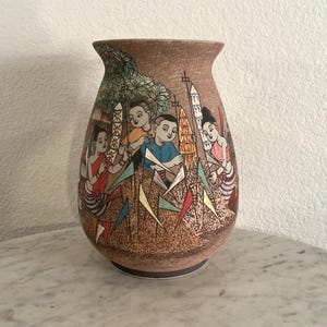 May include: Brown ceramic vase with a detailed illustration of people holding decorative objects. The artwork features figures in colorful clothing, set against a backdrop of trees and flags. The vase has a textured surface and a flared top.