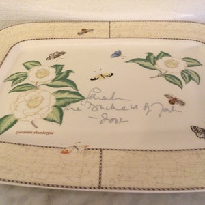 Puede incluir: Un plato rectangular blanco para servir con un borde marr&oacute;n y un dise&ntilde;o floral con flores blancas, hojas verdes y mariposas. El texto "Gardenia thunbergia" y "Sarah The Duchess of York - 2001" est&aacute; impreso en el plato.