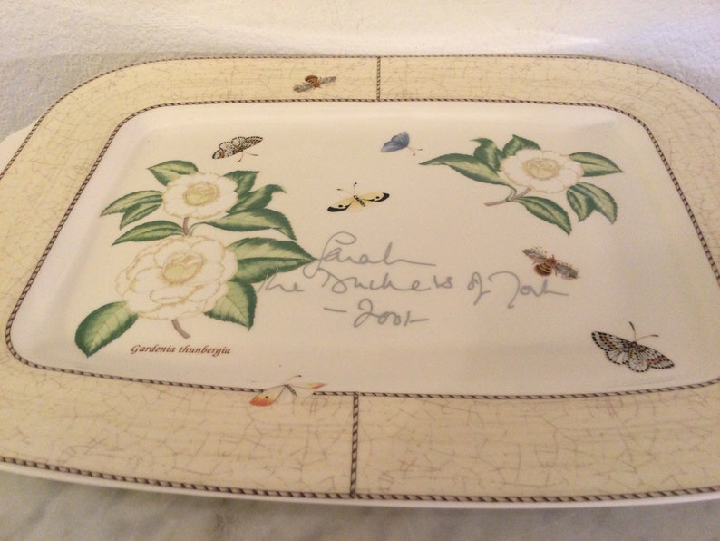 Puede incluir: Un plato rectangular blanco para servir con un borde marr&oacute;n y un dise&ntilde;o floral con flores blancas, hojas verdes y mariposas. El texto "Gardenia thunbergia" y "Sarah The Duchess of York - 2001" est&aacute; impreso en el plato.