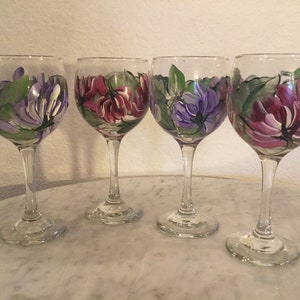 Peut inclure: Quatre verres &agrave; vin peints &agrave; la main avec des motifs floraux. Chaque verre pr&eacute;sente un arrangement diff&eacute;rent de fleurs violettes, roses et vertes.