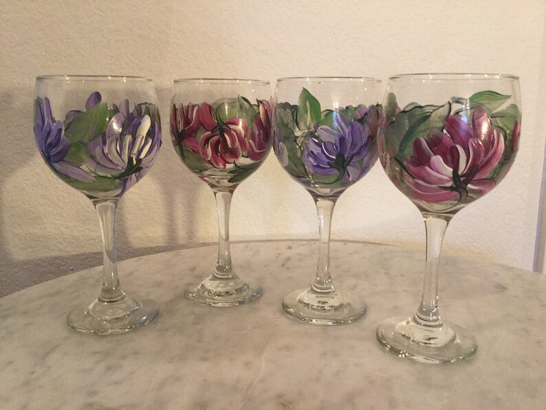 Peut inclure: Quatre verres &agrave; vin peints &agrave; la main avec des motifs floraux. Chaque verre pr&eacute;sente un arrangement diff&eacute;rent de fleurs violettes, roses et vertes.