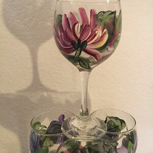 Peut inclure: Trois verres &agrave; vin avec des motifs floraux peints &agrave; la main. Le verre du haut pr&eacute;sente une grande fleur rose avec des feuilles vertes. Les deux verres du bas pr&eacute;sentent des fleurs violettes plus petites avec des feuilles vertes.