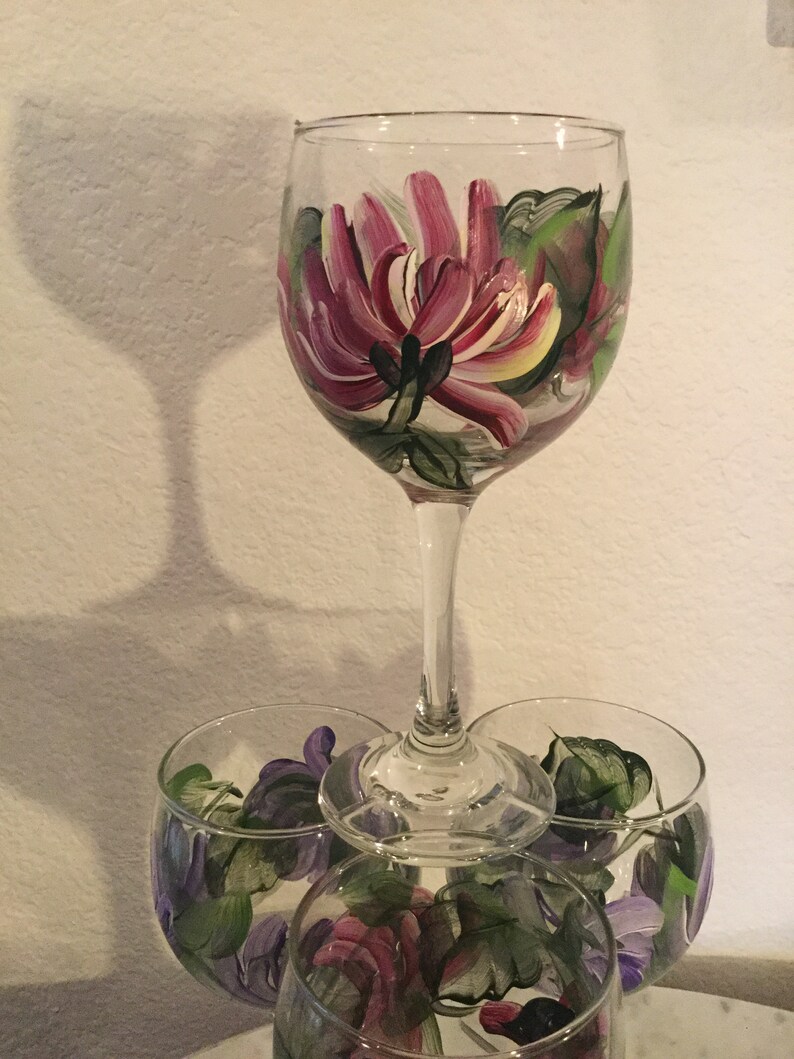 Peut inclure: Trois verres &agrave; vin avec des motifs floraux peints &agrave; la main. Le verre du haut pr&eacute;sente une grande fleur rose avec des feuilles vertes. Les deux verres du bas pr&eacute;sentent des fleurs violettes plus petites avec des feuilles vertes.