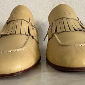 Puede incluir: Un par de mocasines de cuero amarillo claro con flecos. Los zapatos tienen una punta redondeada, costuras decorativas y una suela marr&oacute;n oscuro. Los mocasines se exhiben sobre una superficie de color claro.