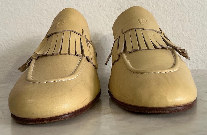 Puede incluir: Un par de mocasines de cuero amarillo claro con flecos. Los zapatos tienen una punta redondeada, costuras decorativas y una suela marr&oacute;n oscuro. Los mocasines se exhiben sobre una superficie de color claro.