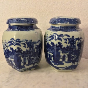 Pode incluir: Dois jarros de porcelana azul e branca com tampas. Os jarros têm forma hexagonal e apresentam um design tradicional de paisagem chinesa.