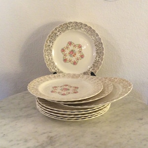 Puede incluir: Una pila de diez platos blancos para cenar con un borde dorado y un diseño floral. Los platos tienen un borde festoneado y están hechos de cerámica.