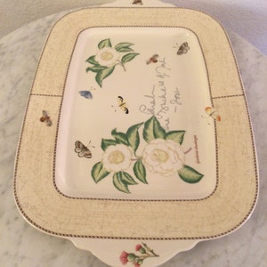 Puede incluir: Una bandeja rectangular para servir con fondo crema y borde marr&oacute;n. La bandeja presenta un dise&ntilde;o floral con camelias blancas, hojas verdes y mariposas. El texto "Cada flor es un punto en la textura de la alegr&iacute;a" est&aacute; impreso en la bandeja.