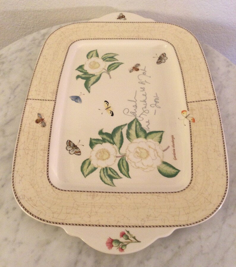 Puede incluir: Una bandeja rectangular para servir con fondo crema y borde marr&oacute;n. La bandeja presenta un dise&ntilde;o floral con camelias blancas, hojas verdes y mariposas. El texto "Cada flor es un punto en la textura de la alegr&iacute;a" est&aacute; impreso en la bandeja.