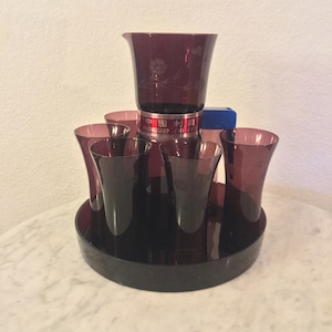 Può includere: Un set di sei bicchieri di vetro viola e una caraffa abbinata con un motivo floreale inciso. La caraffa ha il testo "ZHONG GUO ZHI ZAO" in rosso sul lato. I bicchieri e la caraffa sono su un vassoio nero.