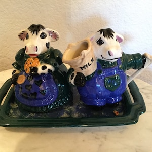 May include: A ceramic cow creamer and sugar set with a green tray. The cow creamer is wearing blue overalls and a green shirt with a bag that says "MILK". The cow sugar dispenser is wearing a green dress with a blue apron and black and white spots.