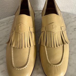 Puede incluir: Un par de mocasines de cuero amarillo claro con flecos. Los zapatos tienen una puntera redondeada y una correa decorativa con un logotipo. Las suelas son de color marr&oacute;n oscuro. Talla 39.