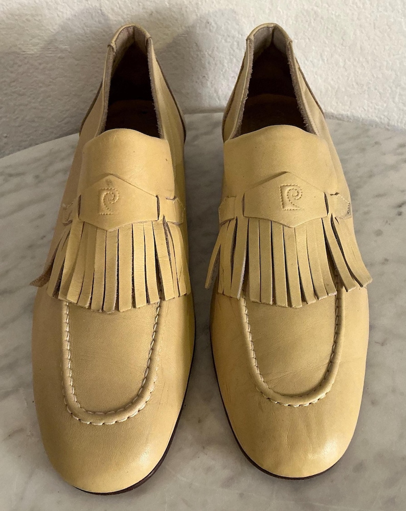 Puede incluir: Un par de mocasines de cuero amarillo claro con flecos. Los zapatos tienen una puntera redondeada y una correa decorativa con un logotipo. Las suelas son de color marr&oacute;n oscuro. Talla 39.