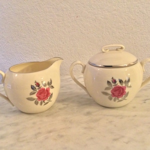 May include: A white creamer and sugar set with a rose floral design. The creamer and sugar are both round and have a single rose with green leaves and pink petals.