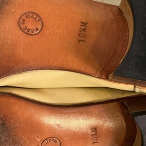Puede incluir: Primer plano de las suelas de dos zapatos de cuero marr&oacute;n. Las suelas est&aacute;n estampadas con "10 1/2 M" y "MADE IN ITALY" dentro de un emblema circular. Los zapatos parecen nuevos.