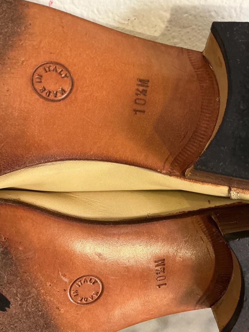 Puede incluir: Primer plano de las suelas de dos zapatos de cuero marr&oacute;n. Las suelas est&aacute;n estampadas con "10 1/2 M" y "MADE IN ITALY" dentro de un emblema circular. Los zapatos parecen nuevos.