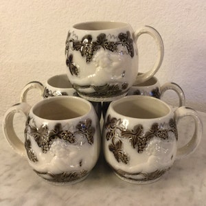 May include: Set of five white ceramic mugs with brown grapevine and fruit designs. Each mug has a rounded body and a handle.