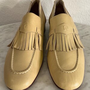 Puede incluir: Un par de mocasines de cuero amarillo claro con flecos y un emblema decorativo. Los zapatos tienen una punta redondeada y un tac&oacute;n bajo. Los mocasines son de estilo cl&aacute;sico.