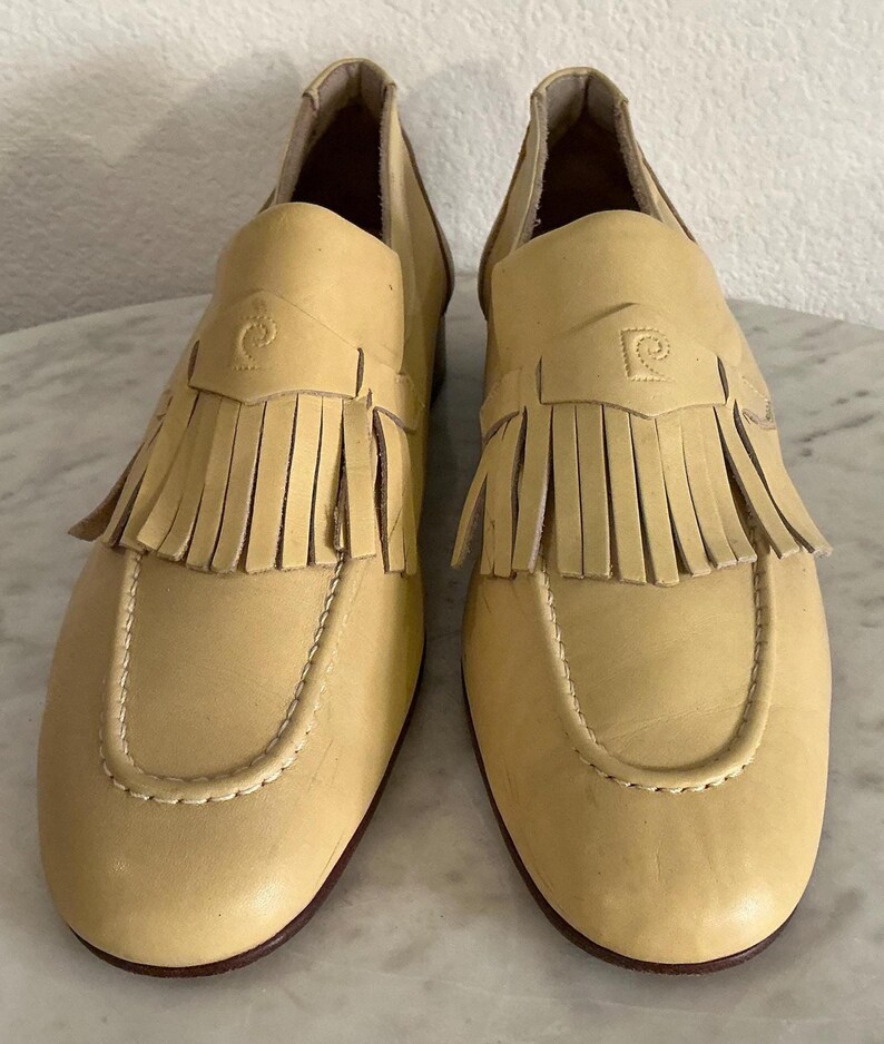 Puede incluir: Un par de mocasines de cuero amarillo claro con flecos y un emblema decorativo. Los zapatos tienen una punta redondeada y un tac&oacute;n bajo. Los mocasines son de estilo cl&aacute;sico.