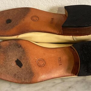 Puede incluir: Un par de zapatos de cuero color crema, con las suelas hacia arriba, con suelas de cuero marr&oacute;n y tacones negros. Las suelas est&aacute;n marcadas con "10M" y tienen un sello circular que dice "Made in Italy".