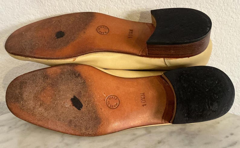 Puede incluir: Un par de zapatos de cuero color crema, con las suelas hacia arriba, con suelas de cuero marr&oacute;n y tacones negros. Las suelas est&aacute;n marcadas con "10M" y tienen un sello circular que dice "Made in Italy".