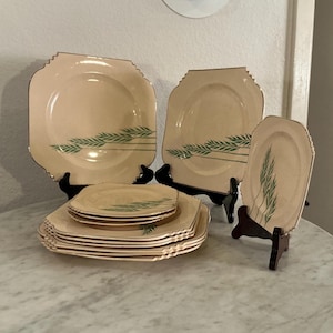 May include: A collection of vintage beige square plates with gold trim and a green wheat stalk design. The set includes various sizes, displayed on black plate stands, showcasing a classic dinnerware set.