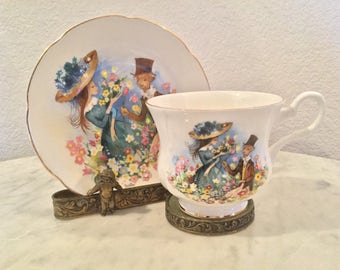 May include: A white teacup and saucer set with a floral design featuring two couples in a garden setting. The teacup has a gold rim and the saucer has a gold rim and a gold band around the edge.