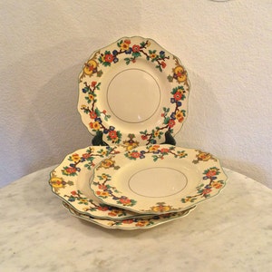 May include: A set of four cream-colored plates with scalloped edges. Each plate features a floral design in red, yellow, blue, and green, with a gold rim. The plates are stacked and displayed on a white surface.