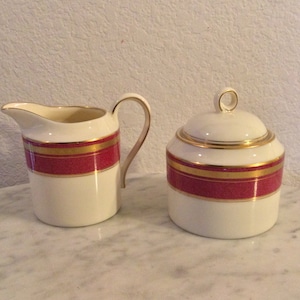 May include: A white creamer and sugar set with a red and gold band design.