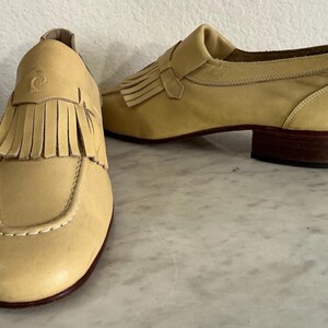 Puede incluir: Un par de mocasines de cuero amarillo claro con flecos. Los zapatos tienen una puntera redondeada, un tac&oacute;n bajo y una correa decorativa. El logotipo de Pierre Cardin es visible en el lateral de un zapato.